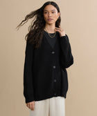 A woman with long wavy hair wears the Cashmere Cocoon Cardigan in black over a black top and cream pants, accessorized with gold jewelry. She stands against a beige background, gazing slightly to the side with one hand near her neck.