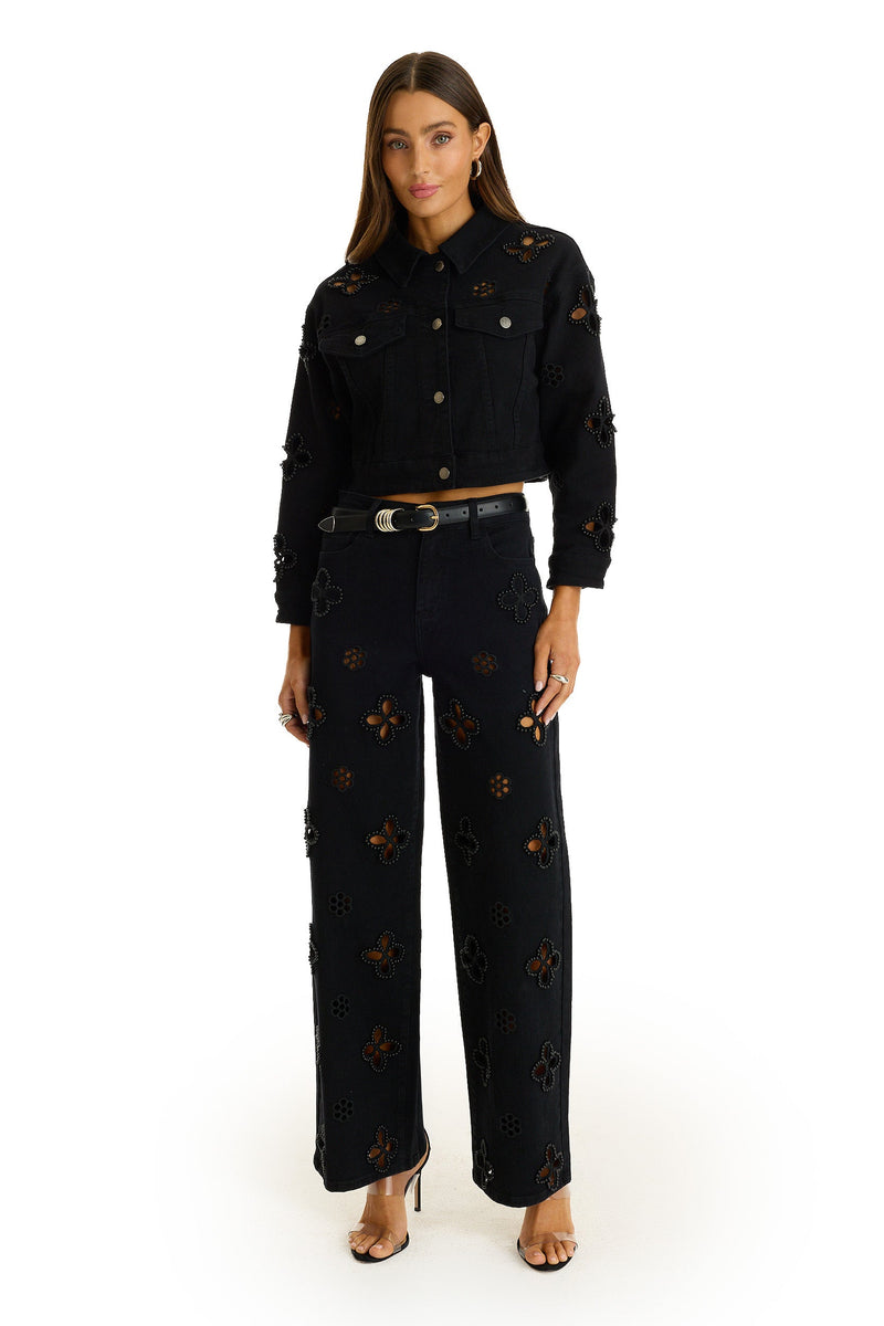 A woman stands facing forward in the Kaia Pearl High Rise Jean - Black, featuring cut-out floral patterns and pearl embroidery, paired with a matching black jacket, gold-buckle belt, and open-toe heels against a white background.