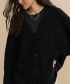 A woman models the Cashmere Cocoon Cardigan in black over a matching top, styled with layered gold necklaces. She poses with one hand on her hip, her long wavy dark hair contrasting against a beige background.