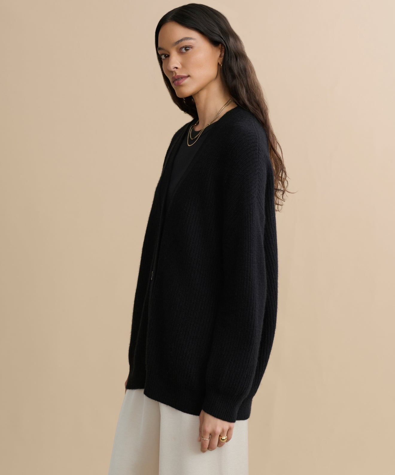 A woman with long dark hair, wearing the Cashmere Cocoon Cardigan in black, cream pants, and simple gold jewelry, stands against a plain beige background, looking slightly to the side.