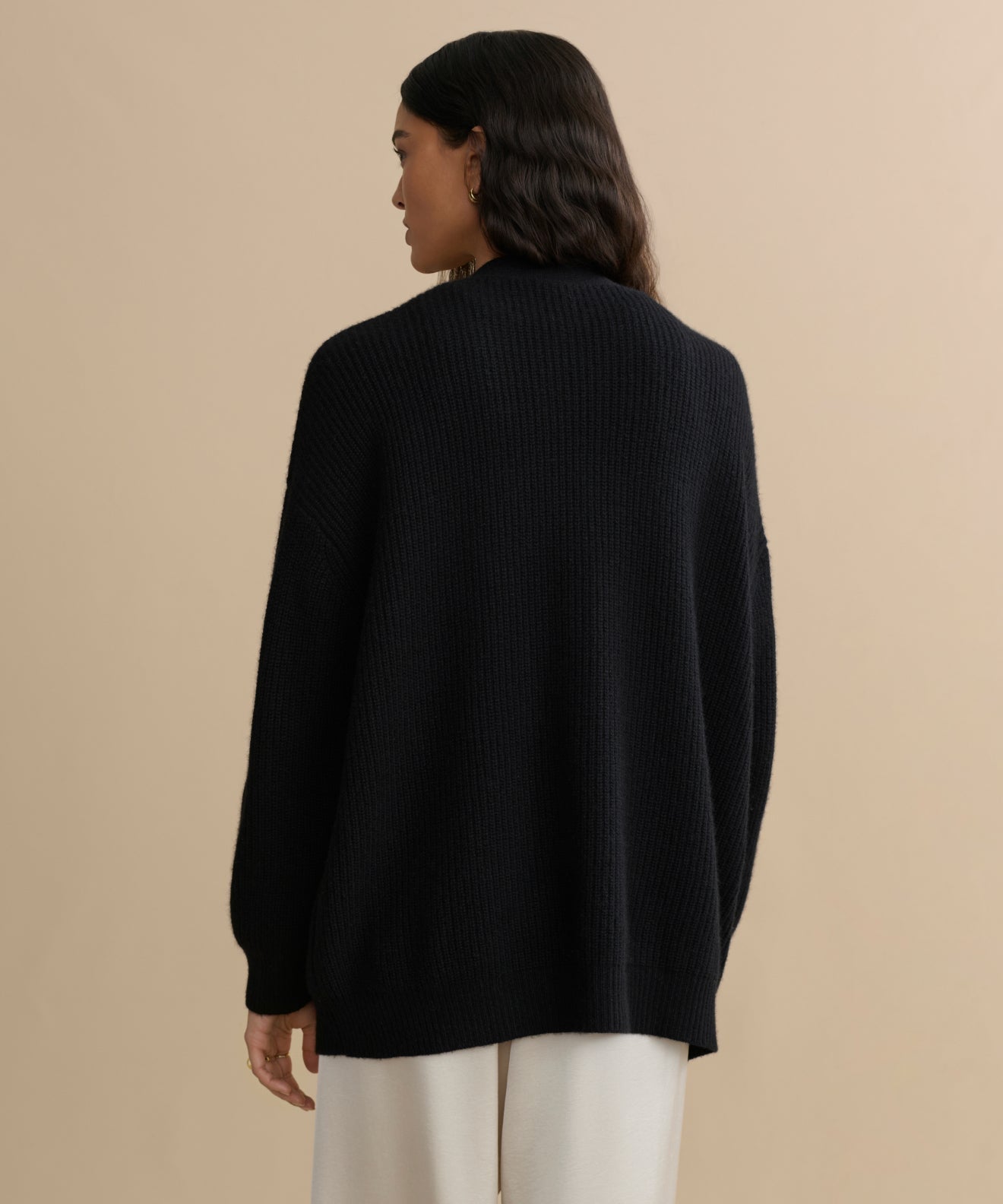 A person with long dark hair, wearing the oversized black Cashmere Cocoon Cardigan and light-colored pants, stands facing away against a plain beige background.