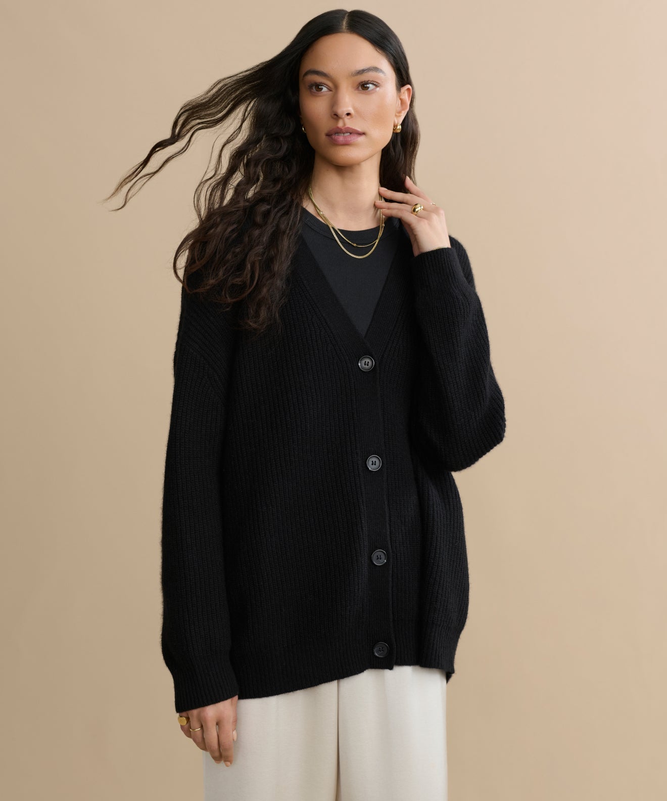 A woman with long wavy hair wears the Cashmere Cocoon Cardigan in black over a black top and cream pants, accessorized with gold jewelry. She stands against a beige background, gazing slightly to the side with one hand near her neck.